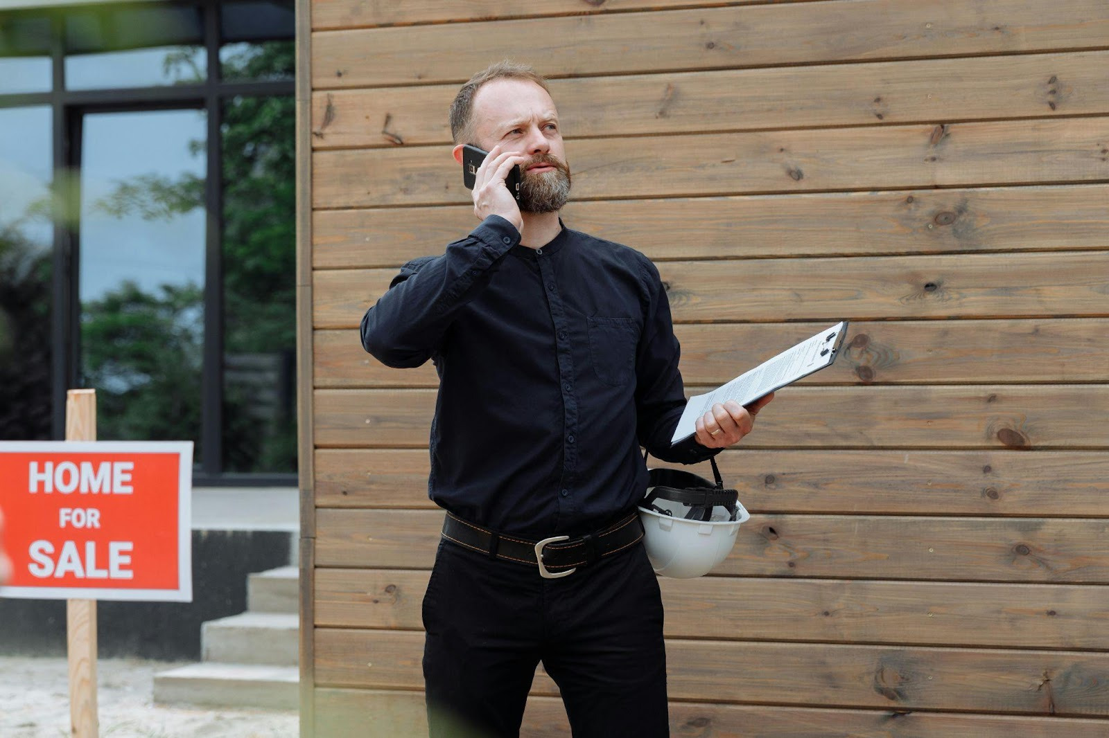 A man in black clothing holds a phone to his ear and a clipboard, standing near a wooden wall. A 