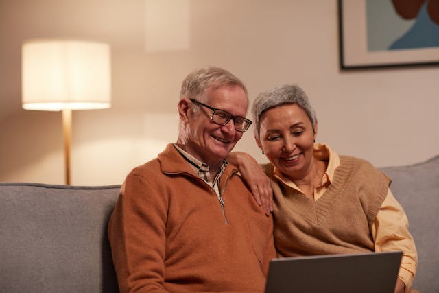 An elderly couple sits on a sofa, smiling while looking at a laptop. The room is warmly lit, creating a cozy atmosphere.